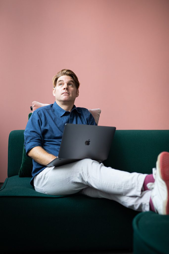 Porträt-rothaariger-mann-auf-Sofa-mit-laptop-auf-schoß-vor-rosa-hintergrund