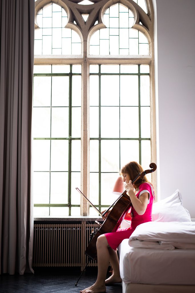 Porträt-einer-rothaarigen-Cellistin-mit-Cello-auf-Hotelbett-sitzend-vor-Fenster-im-Qvest-Hotel